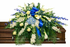 Elegant casket spray featuring white roses, white calla lilies, blue hydrangea, delicate blue delphiniums , and lush greenery, accented with a royal blue satin bow and arranged atop a wooden casket in a softly lit chapel setting.