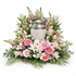 Graceful memorial urn arrangement featuring a silver urn surrounded by soft pink roses, pink gerbera daisies, white lilies, snapdragons, and lush greenery, designed in a full, elevated style on a wooden pedestal against a clean white background.
