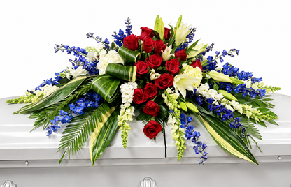 Patriotic casket spray featuring deep red roses, white stock, white lilies and white filler flowers, vibrant blue delphinium, and cascading greenery, arranged in a bold, symmetrical design atop a white casket in a softly lit chapel setting.