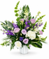 A tall pedestal sympathy arrangement featuring lavender roses, white roses, white hydrangea, white snapdragons, purple stock, Bells of Ireland, lisianthus, and lush tropical greenery arranged in a white pedestal vase. The garden-inspired design showcases varying heights and soft purple, white, and green tones, creating an elegant and peaceful floral tribute for funeral and memorial services.
