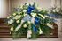 Elegant casket spray featuring white roses, white calla lilies, blue hydrangea, delicate blue delphiniums , and lush greenery, accented with a royal blue satin bow and arranged atop a wooden casket in a softly lit chapel setting.