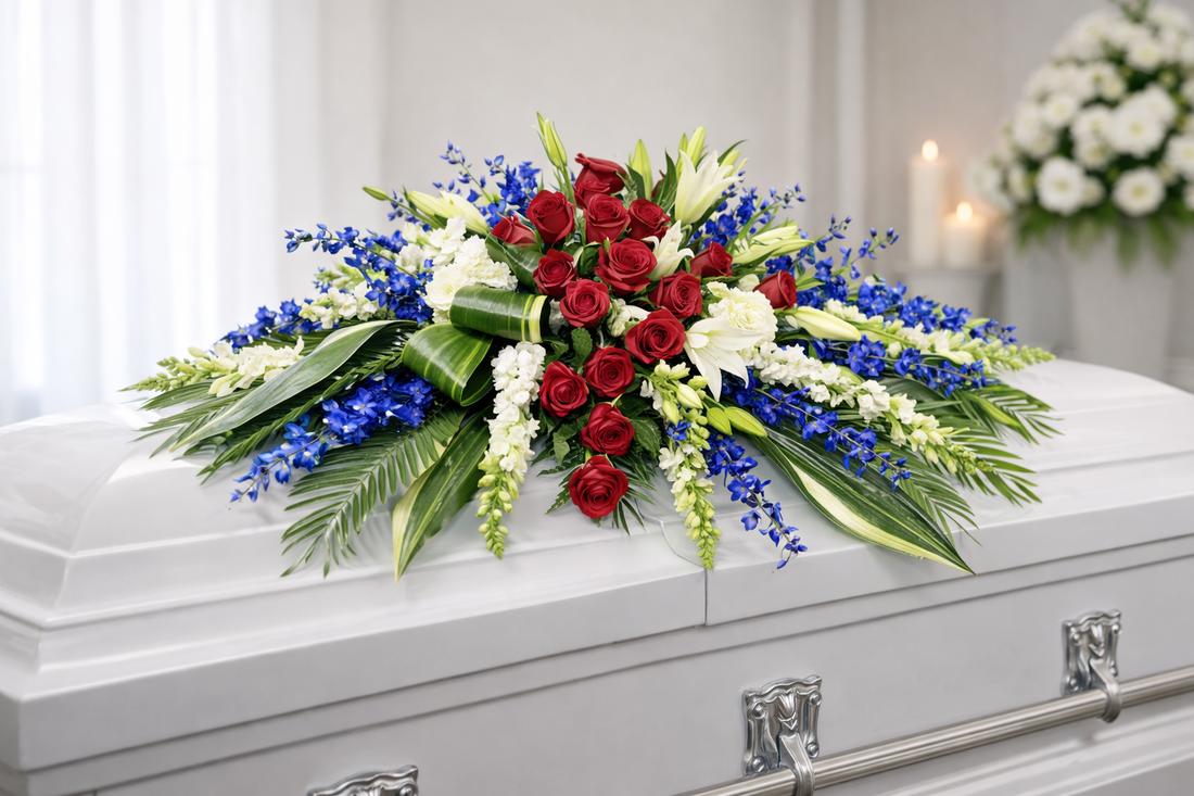 Patriotic casket spray featuring deep red roses, white stock, white lilies and white filler flowers, vibrant blue delphinium, and cascading greenery, arranged in a bold, symmetrical design atop a white casket in a softly lit chapel setting.