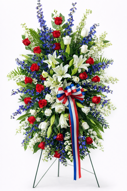 Patriotic standing sympathy spray featuring red roses, white lilies, blue delphinium, and white filler flowers, accented with lush greenery and a red, white, and blue ribbon, arranged in a tall, symmetrical design on a green easel against a clean white background.