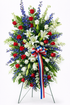 Patriotic standing sympathy spray featuring red roses, white lilies, blue delphinium, and white filler flowers, accented with lush greenery and a red, white, and blue ribbon, arranged in a tall, symmetrical design on a green easel against a clean white background.