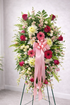 Elegant standing sympathy spray featuring pink and red roses, pink gerbera daisies, white chrysanthemums, soft filler flowers, and lush greenery, accented with a flowing pink satin ribbon and displayed on a green easel against a neutral background.