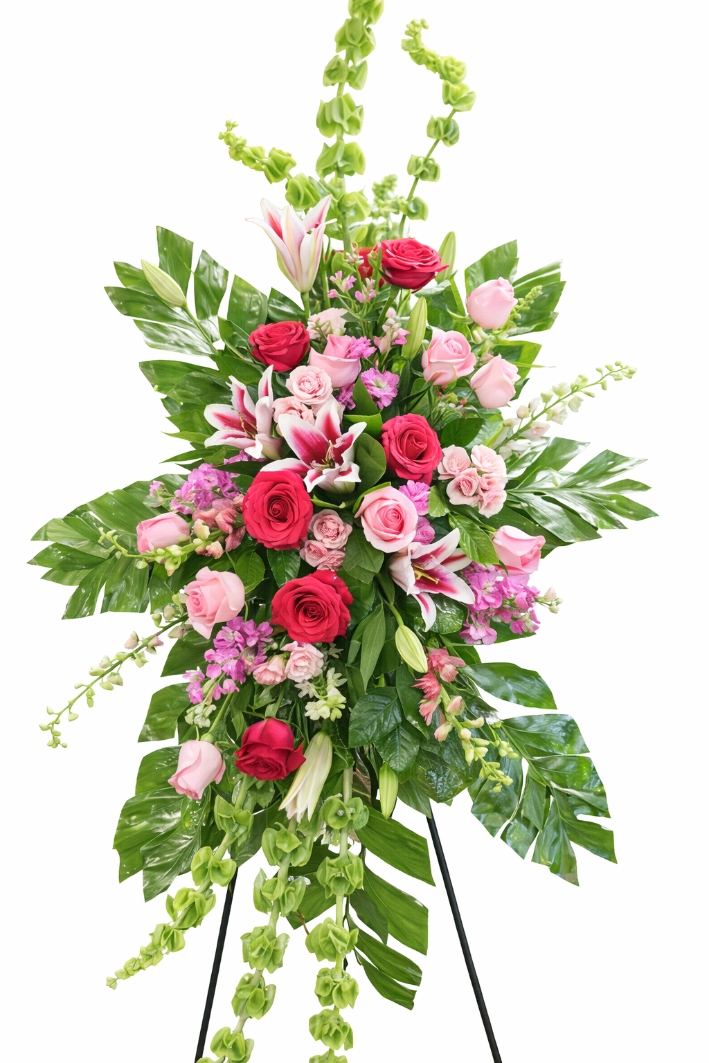 Vibrant standing sympathy spray featuring red and pink roses, pink lilies, bells of Ireland, soft pink filler flowers, and cascading palms, arranged in a lush vertical design on a green easel against a clean white background.