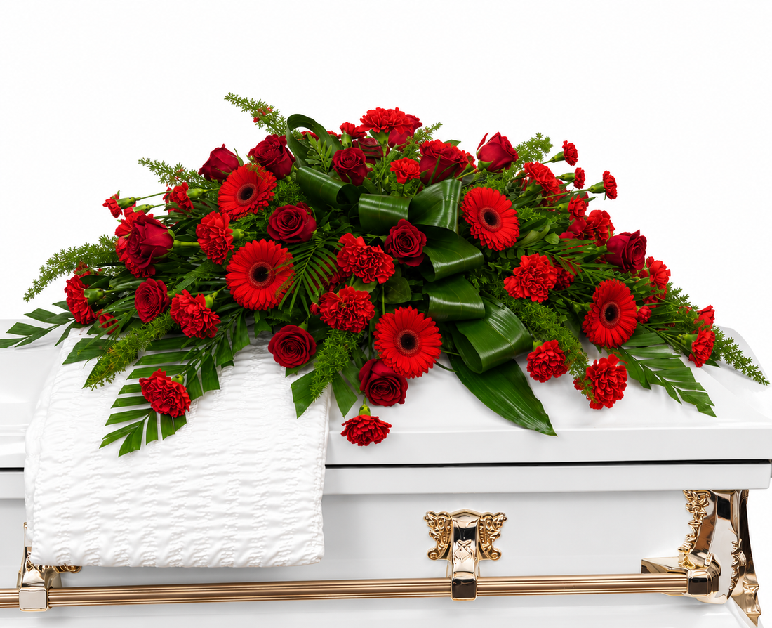 A large red and green floral casket bouquet with red roses, gerbera daisies, carnations, and green foliage, arranged on a white casket with gold hardware and a folded white cloth.