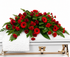 A large red and green floral casket bouquet with red roses, gerbera daisies, carnations, and green foliage, arranged on a white casket with gold hardware and a folded white cloth.