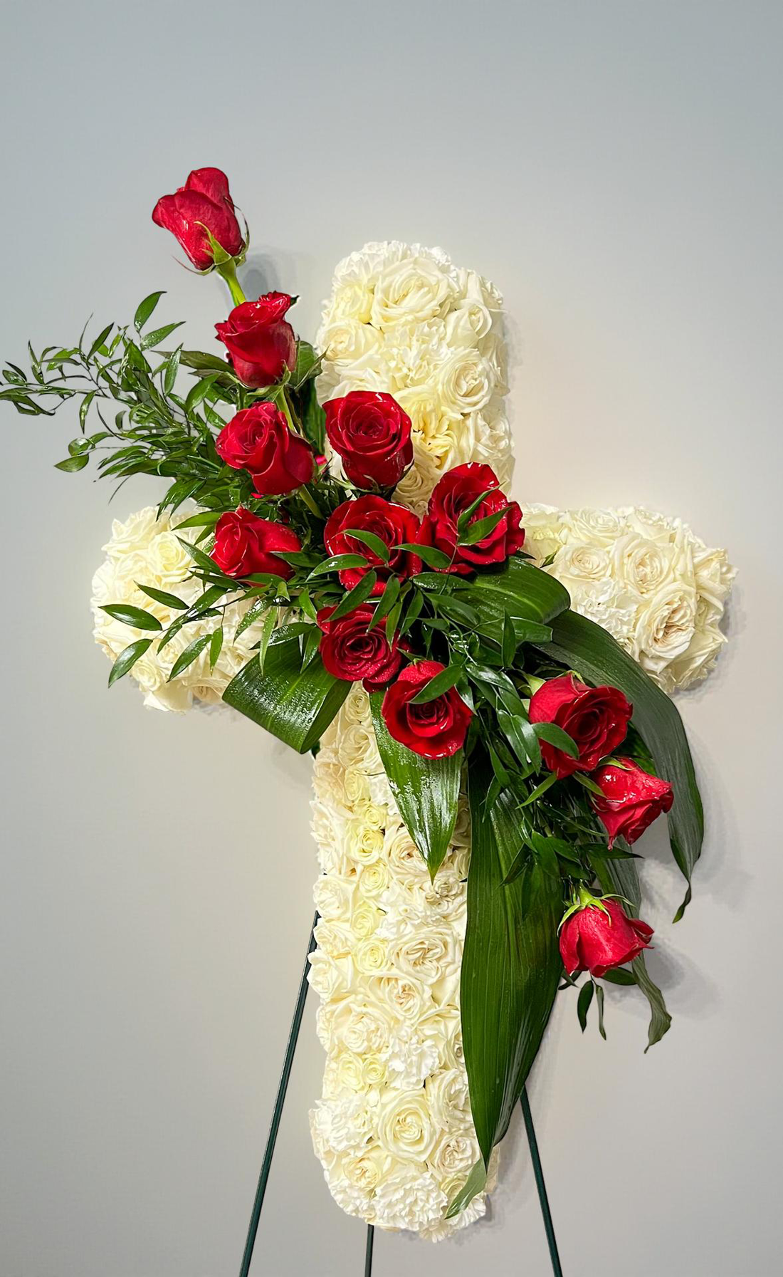 Cross-shaped sympathy arrangement made of ivory roses and white hydrangeas, accented with cascading red roses and lush green foliage, designed in a graceful, traditional style and displayed on a green easel against a white background.