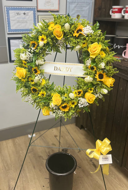 A yellow and sunflower memorial flower wreath on a stand, with a central sign reading &