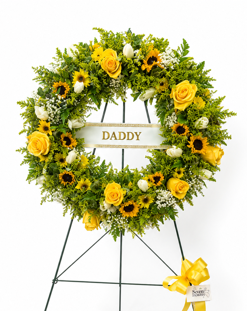 A circular floral wreath on a black metal stand, featuring yellow roses, sunflowers, white tulips, and green foliage, with a cream banner in the center reading &