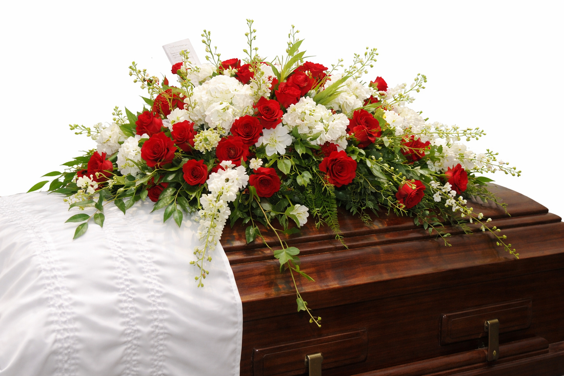 Elegant casket spray featuring red roses, white lilies, white hydrangea, delicate white daisies, and cascading greenery, arranged in a full, symmetrical design atop a polished wood casket in a softly lit chapel setting.