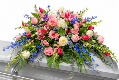 A casket spray featuring pink roses, blue delphinium, white hydrangea, and mixed greenery arranged in a full design across a white casket, displayed against a clean white background.