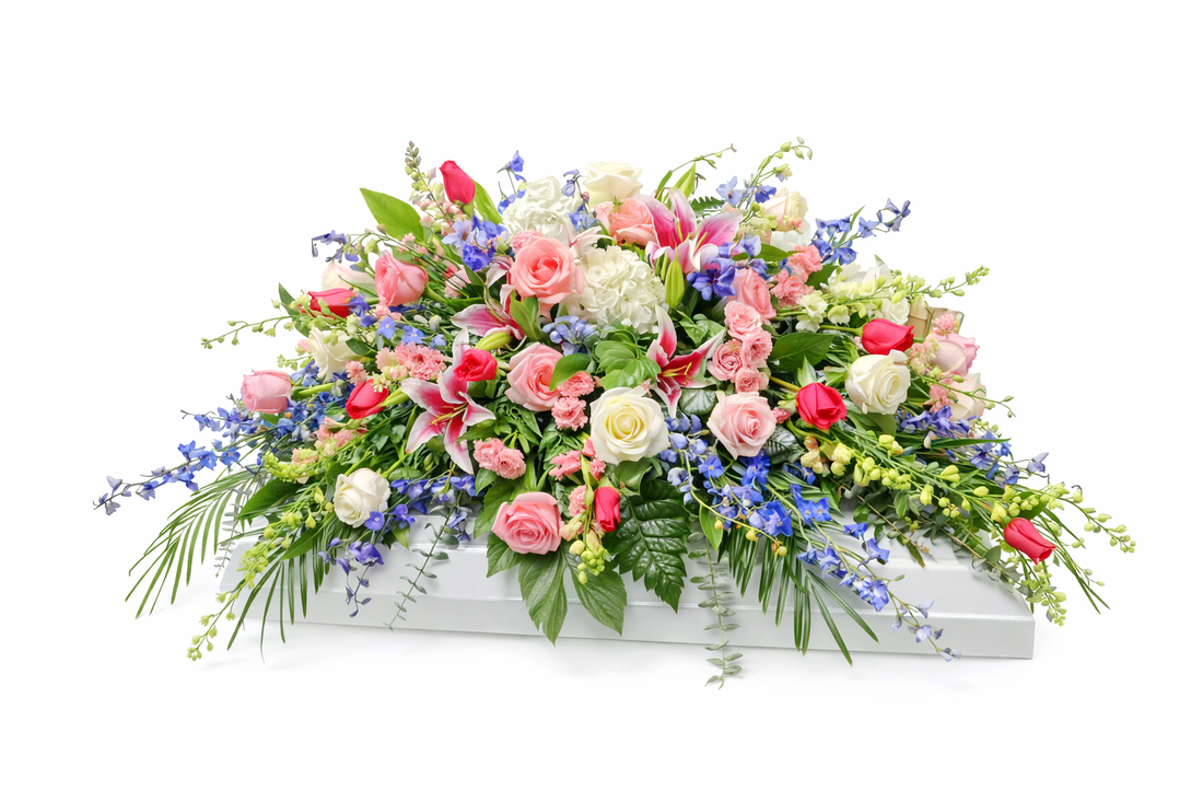 A casket spray featuring pink roses, blue delphinium, white hydrangea, and mixed greenery arranged in a full design across a white casket, displayed against a clean white background.