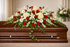 Elegant casket spray featuring red roses, white lilies, white hydrangea, delicate white daisies, and cascading greenery, arranged in a full, symmetrical design atop a polished wood casket in a softly lit chapel setting.