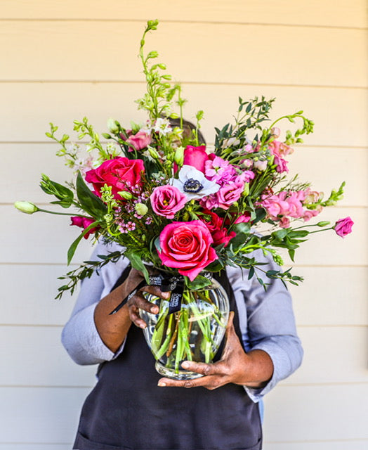 Handheld garden-style floral arrangement featuring bright pink roses, pink lisianthus, white anemone, and mixed greenery in a clear glass vase, photographed outdoors against a light neutral wall.
