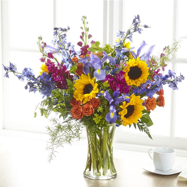 A vibrant bouquet of summer blooms including spray roses, delphiniums, irises, and sunflowers in a clear vase, placed on a table with a cup. Has more flowers than the standard.