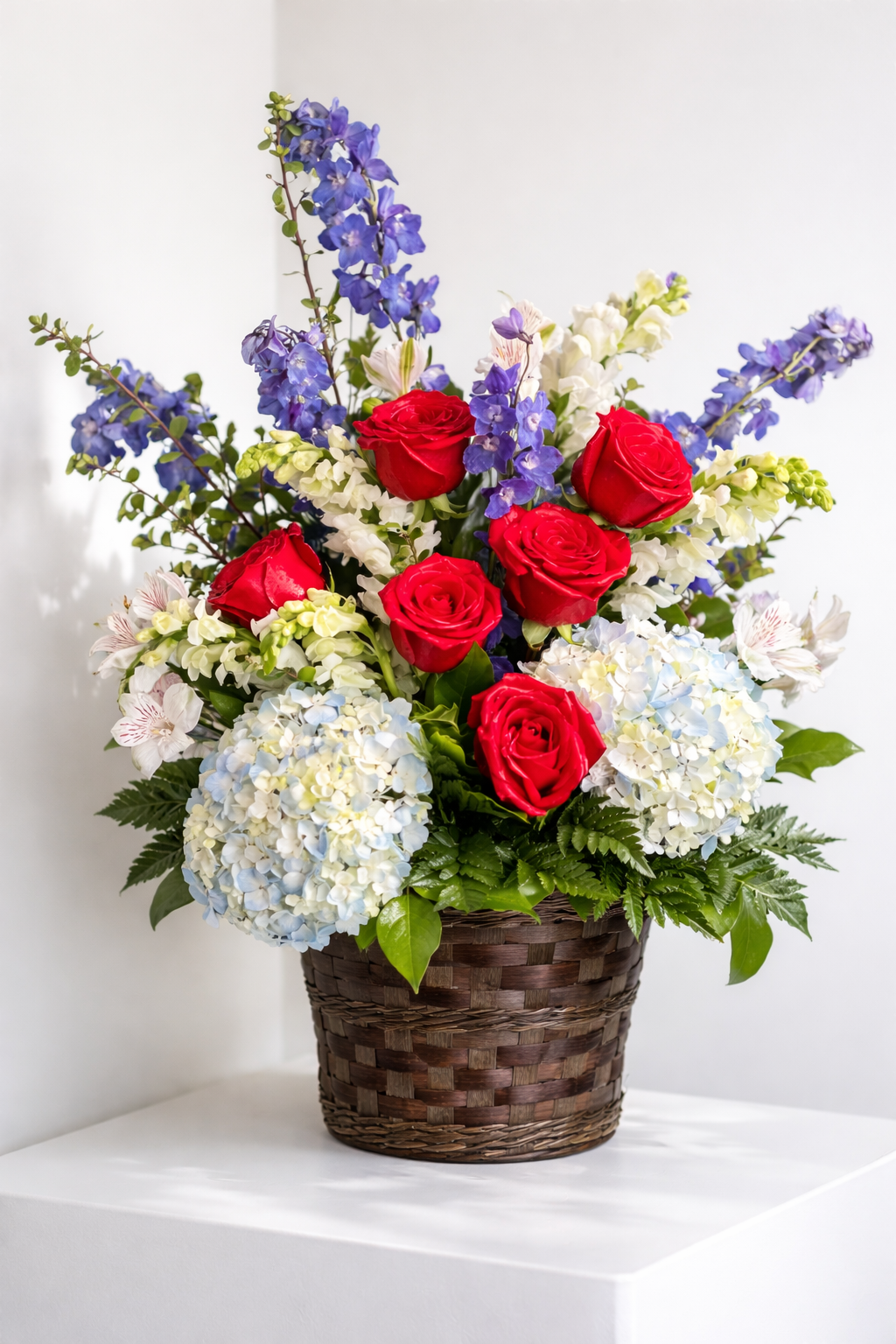 Elegant sympathy basket arrangement featuring red roses, soft blue hydrangea, blue delphinium, white snapdragons, and delicate white accent flowers, complemented by lush green foliage and arranged in a woven basket against a clean white background.