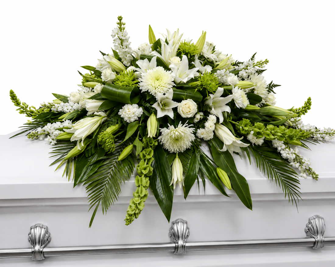 Serene white casket spray featuring white lilies, white stock, white chrysanthemums, white snapdragons, bells of ireland, delicate white filler flowers, and lush cascading greenery, arranged in a full, elegant design atop a white casket in a softly lit chapel setting.