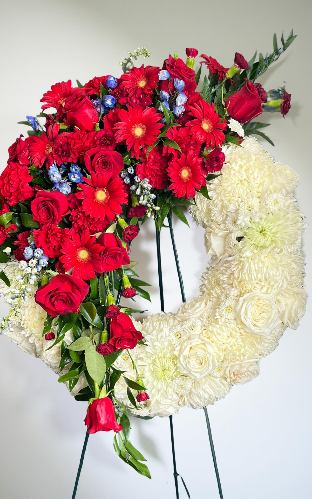 Open-circle sympathy wreath featuring creamy white roses and chrysanthemums, accented with cascading red roses, red gerbera daisies, touches of soft blue delphinium flowers, and lush greenery, arranged in an asymmetrical design on a green easel against a white background.