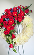 Open-circle sympathy wreath featuring creamy white roses and chrysanthemums, accented with cascading red roses, red gerbera daisies, touches of soft blue delphinium flowers, and lush greenery, arranged in an asymmetrical design on a green easel against a white background.