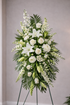 Classic white standing sympathy spray featuring white lilies, white roses, white cremon mums, carnations, delicate filler flowers, and lush palm and fern greenery, arranged in a full, cascading design on a green easel against a soft neutral background.