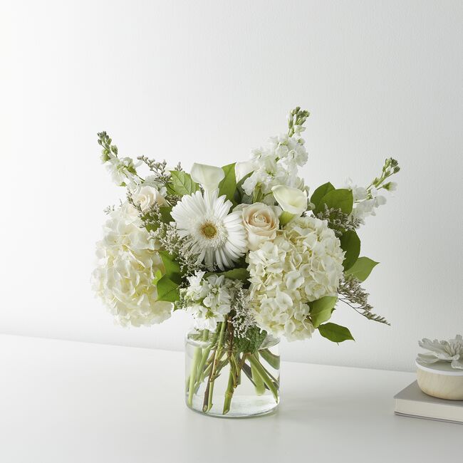 A beautiful bouquet of white roses, hydrangeas, gerbera daisies, mini calla lilies, and Limonium arranged in a clear glass vase. The background is a clean, minimalist white backdrop with soft natural lighting, featuring a smooth white wall and a simple tabletop surface. Subtle neutral décor elements, including a small, stacked book and a decorative accent, sit off to the side, creating a calm, modern setting that keeps the focus on the floral arrangement.