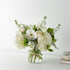 A beautiful bouquet of white roses, hydrangeas, gerbera daisies, mini calla lilies, and Limonium arranged in a clear glass vase. The background is a clean, minimalist white backdrop with soft natural lighting, featuring a smooth white wall and a simple tabletop surface. Subtle neutral décor elements, including a small, stacked book and a decorative accent, sit off to the side, creating a calm, modern setting that keeps the focus on the floral arrangement.