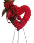 Artificial bleeding heart standing funeral spray made with red silk carnations, cascading red silk flowers, greenery, burgundy satin ribbon, and displayed on a black metal easel.
