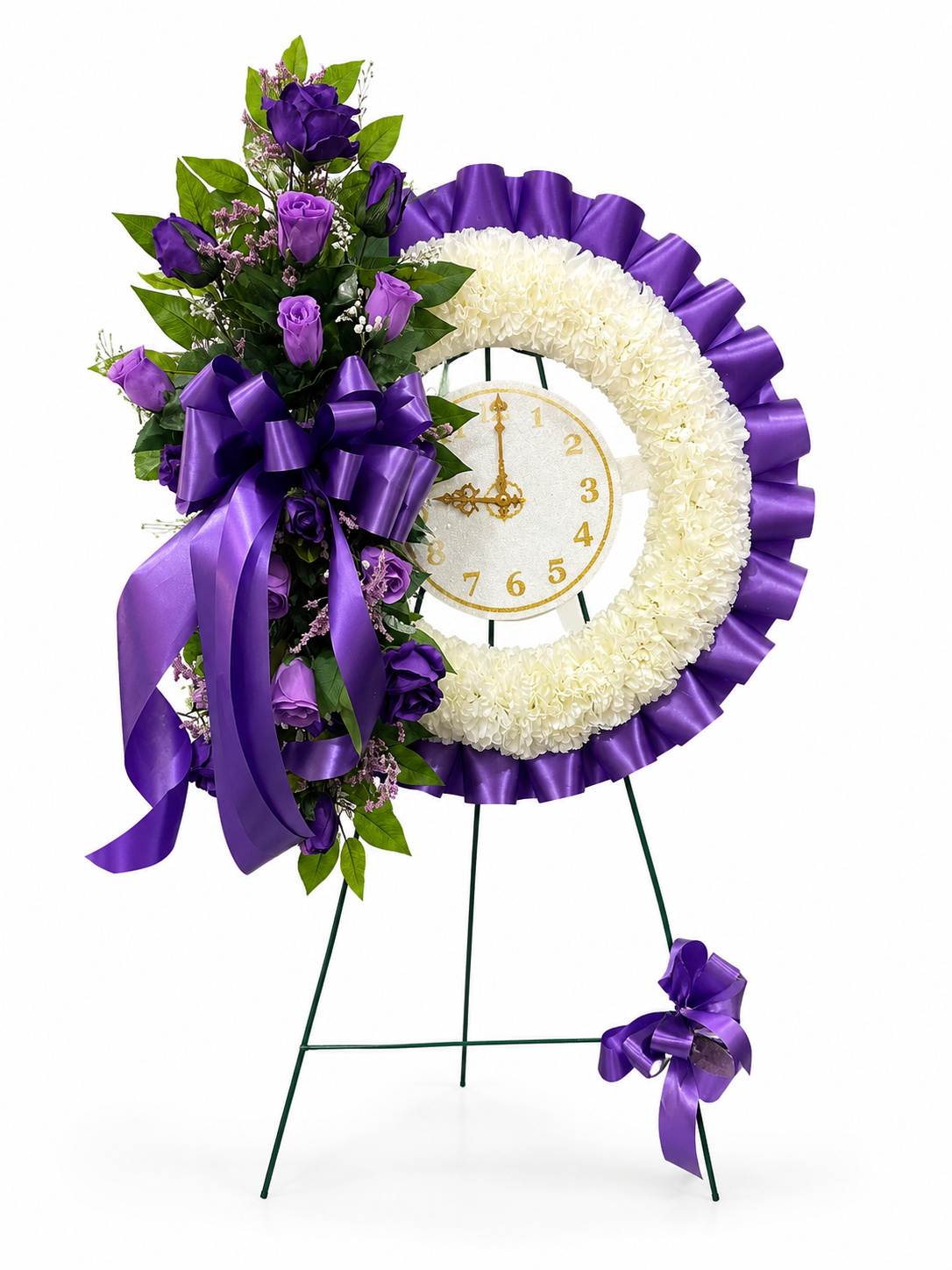 A memorial flower wreath on a metal easel stand, featuring purple roses, white carnations, a purple ruffled border, a white clock face with gold hands, and large purple ribbons.