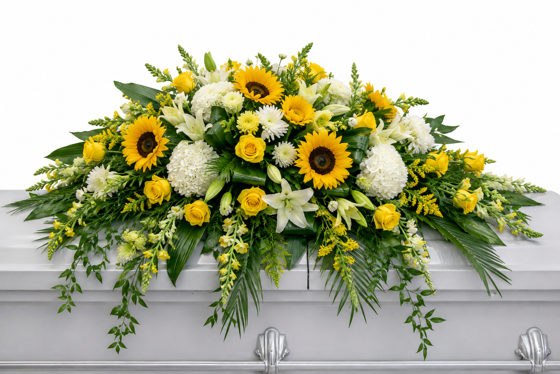 Bright casket spray featuring yellow roses, white hydrangea, sunflowers, white lilies, and cascading greenery, arranged in a full, elegant design atop a white casket in a softly lit chapel setting.