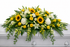 Bright casket spray featuring yellow roses, white hydrangea, sunflowers, white lilies, and cascading greenery, arranged in a full, elegant design atop a white casket in a softly lit chapel setting.