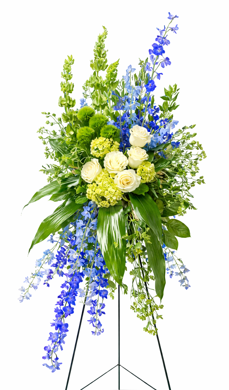 Graceful standing sympathy spray featuring white roses, green hydrangea, vibrant blue delphinium, textured green accents, and cascading greenery, arranged in an elegant vertical design on a green easel against a soft, neutral background.