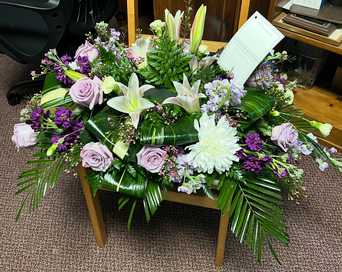 A floral arrangement of white lilies, purple roses, and green foliage displayed on a wooden chair in an indoor room with carpet and furniture visible.