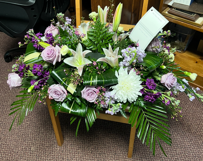 A floral arrangement of white lilies, purple roses, and green foliage displayed on a wooden chair in an indoor room with carpet and furniture visible.