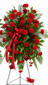 Standing sympathy spray featuring vibrant red carnations, gerbera daisies, and roses accented with lush evergreen foliage, finished with a flowing red satin ribbon, arranged in a classic vertical design on a green easel against a white background.