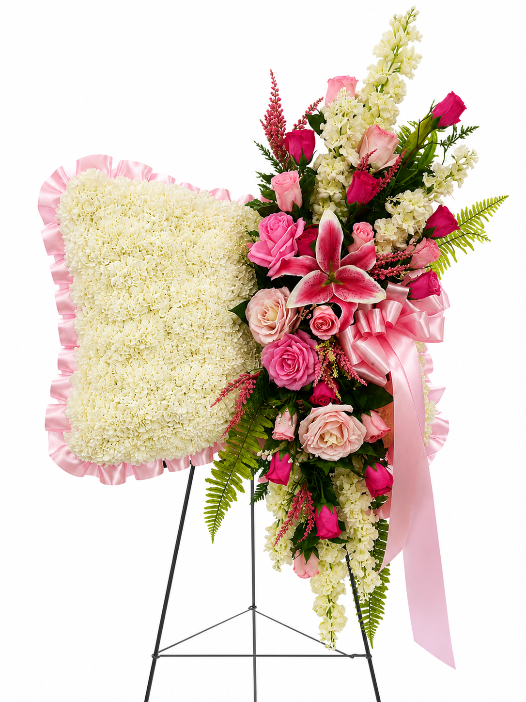 A large floral arrangement on a black metal stand, featuring a square white flower pillow with pink ruffled trim and a cascading bouquet of pink roses, lilies, white flowers, green foliage, and a pink ribbon bow.