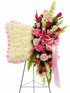A large floral arrangement on a black metal stand, featuring a square white flower pillow with pink ruffled trim and a cascading bouquet of pink roses, lilies, white flowers, green foliage, and a pink ribbon bow.