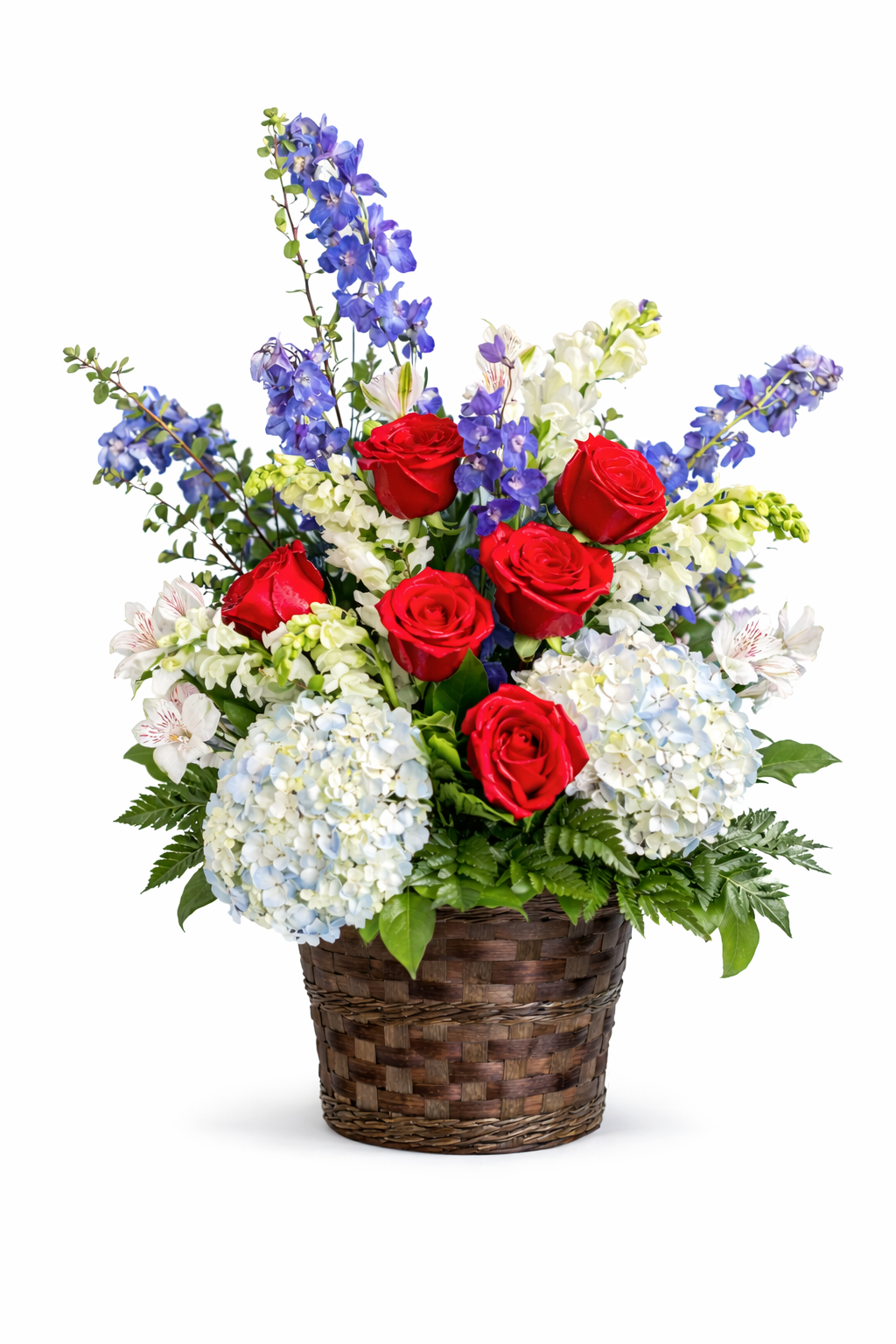 Elegant sympathy basket arrangement featuring red roses, soft blue hydrangea, blue delphinium, white snapdragons, and delicate white accent flowers, complemented by lush green foliage and arranged in a woven basket against a clean white background.