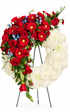Open-circle sympathy wreath featuring creamy white roses and chrysanthemums, accented with cascading red roses, red gerbera daisies, touches of soft blue delphinium flowers, and lush greenery, arranged in an asymmetrical design on a green easel against a white background.